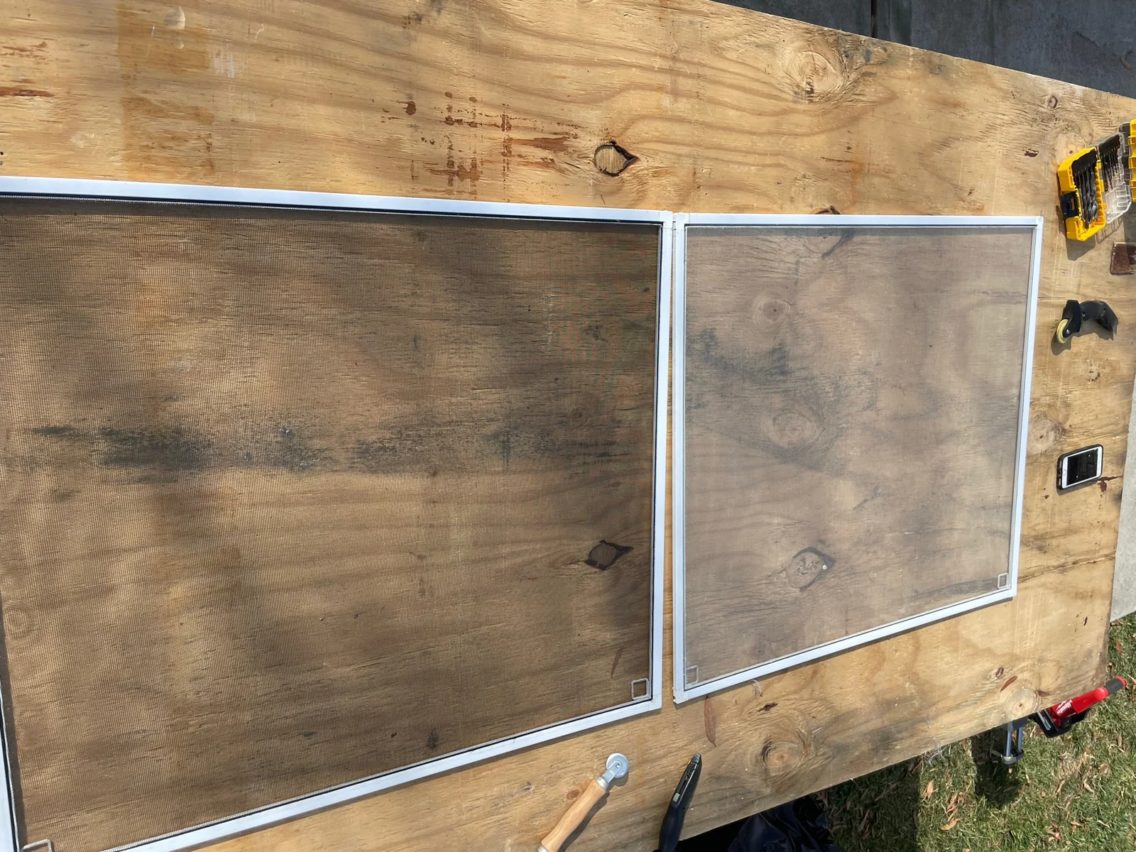Two white-framed window screens on a worktable, with newer dark charcoal mesh on the left and worn whitening fiberglass mesh on the right.